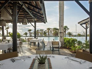 A restaurant with tables set for dining, overlooking a pool and palm trees.