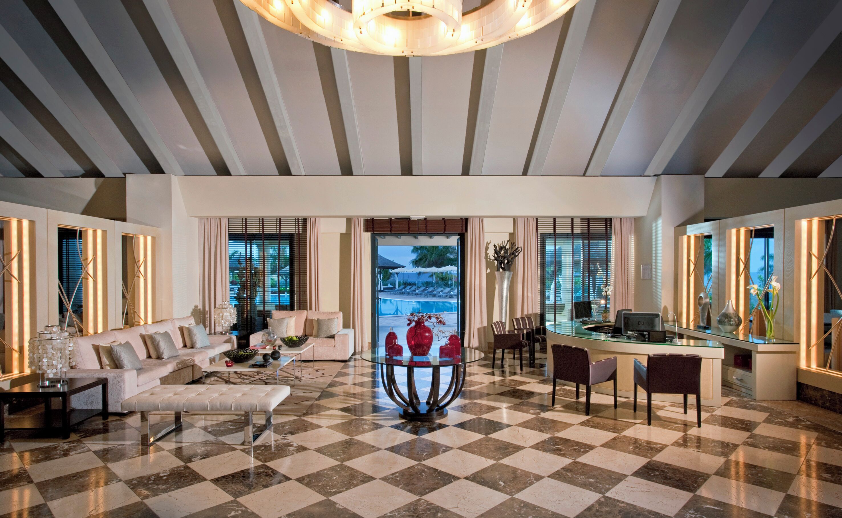 A spacious hotel lobby with a checkered floor, a central seating area with a sofa and armchairs, a glass-top coffee table, and a view of an outdoor pool area through large windows.