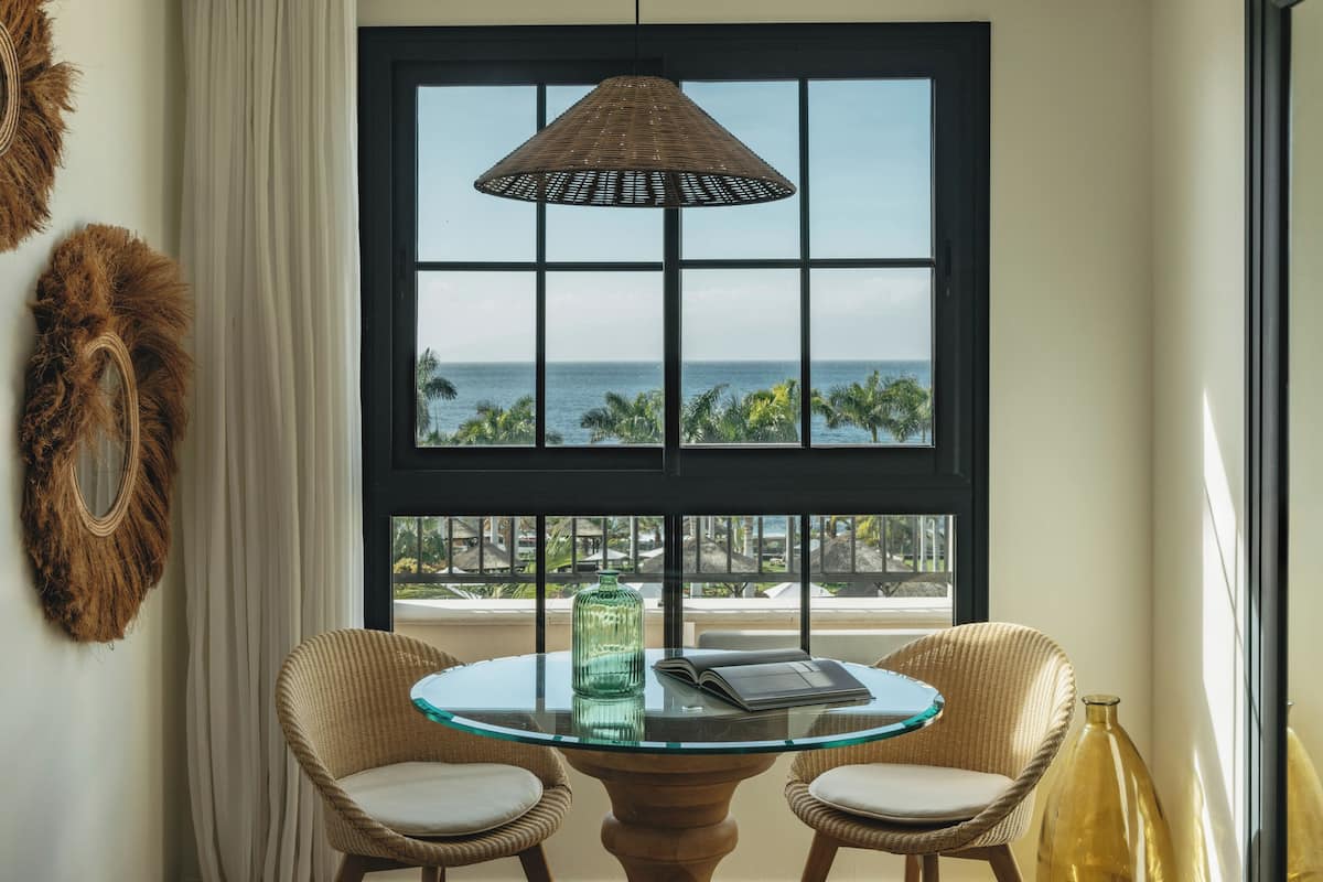 Une pièce avec une table en verre, deux chaises en osier, une grande fenêtre donnant sur l’océan et une lampe suspendue.