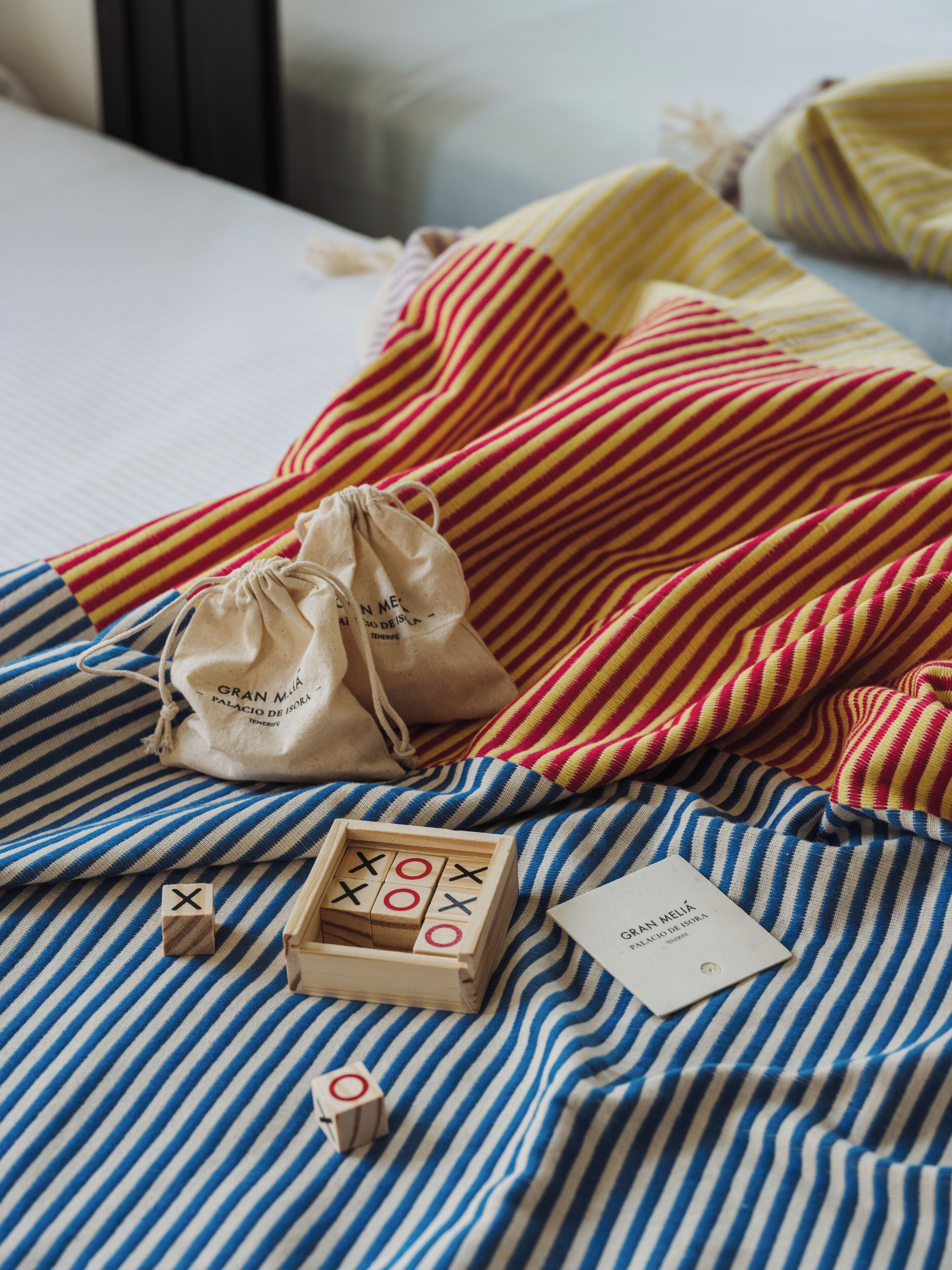 A bed with striped bedding, a small wooden tic-tac-toe game, a drawstring bag, and a tag with text.