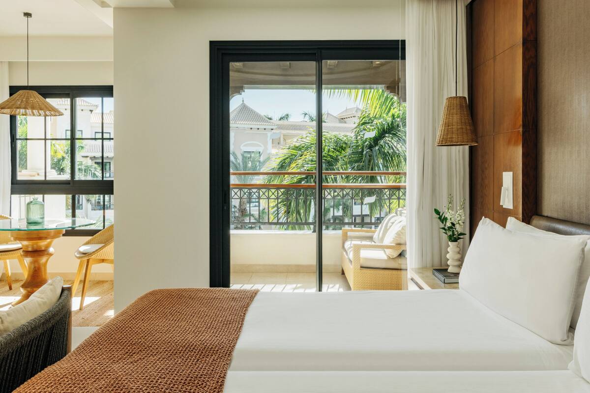 Une chambre d’hôtel avec un lit, un balcon donnant sur des palmiers, un coin repas avec une table en verre et des meubles en osier.