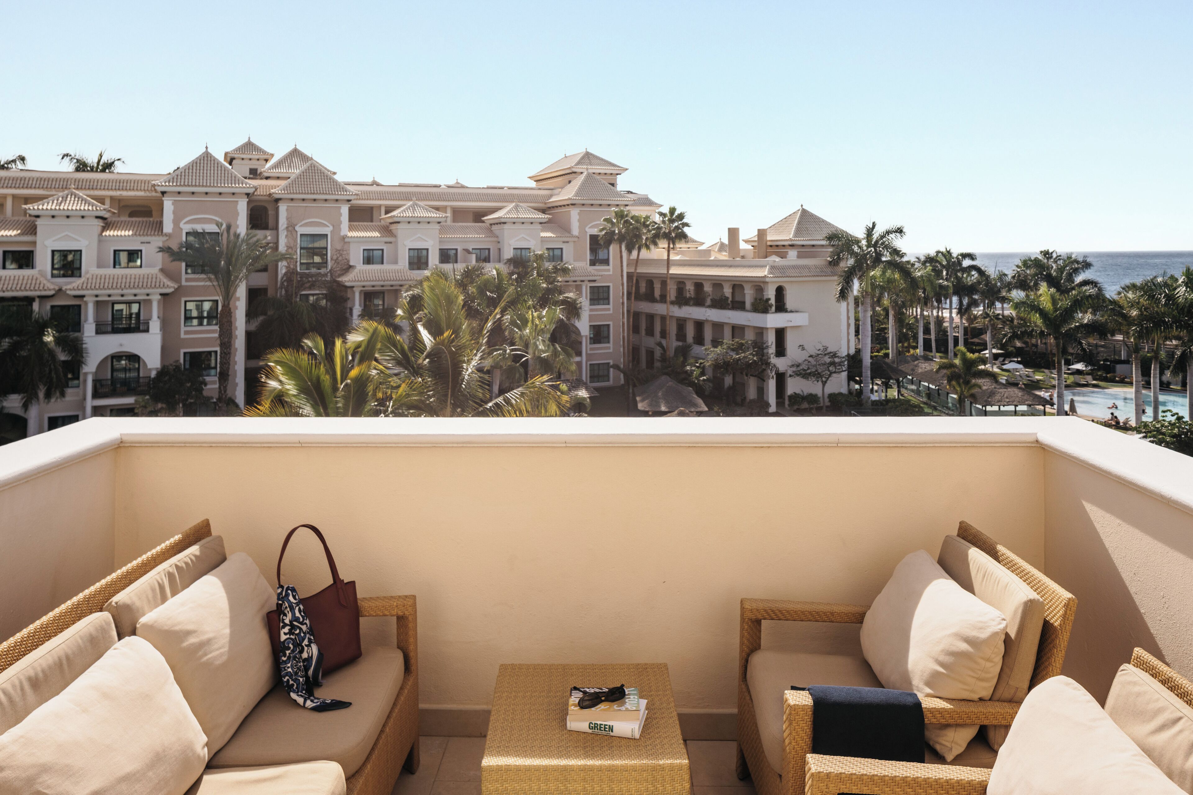 Un balcón con mobiliario de mimbre, una mesita y vistas a un edificio de resort con palmeras.