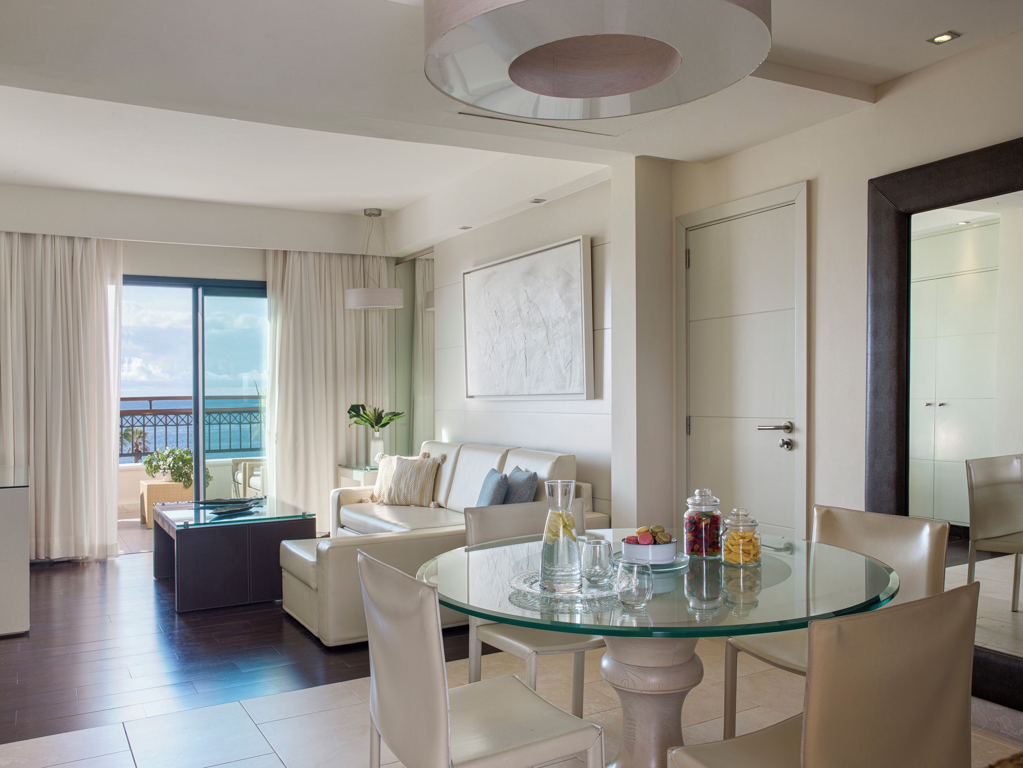 A modern living room with a glass dining table, white leather sofas, and a view of the ocean.