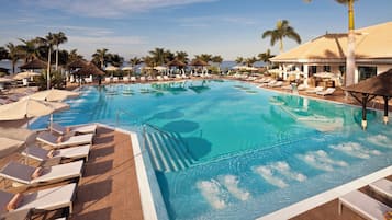 Ein großer Swimmingpool mit Liegestühlen und Sonnenschirmen, umgeben von Palmen und einem Gebäude mit einem Ziegeldach.