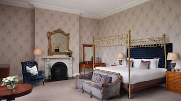 A bedroom with a four-poster bed, a fireplace, a blue armchair, a sofa, and a chandelier.