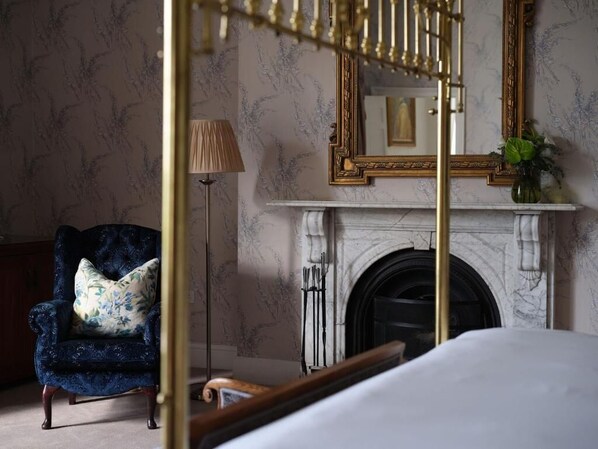 A bedroom with a four-poster bed, a blue armchair with a floral pillow, a standing lamp, and a fireplace with a marble mantle. - Lyrath Estate Hotel Spa & Convention Centre (Kilkenny)