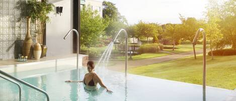 Una piscina interior moderna con paredes de vidrio transparente, vistas a un jardín cuidado y una fuente integrada.