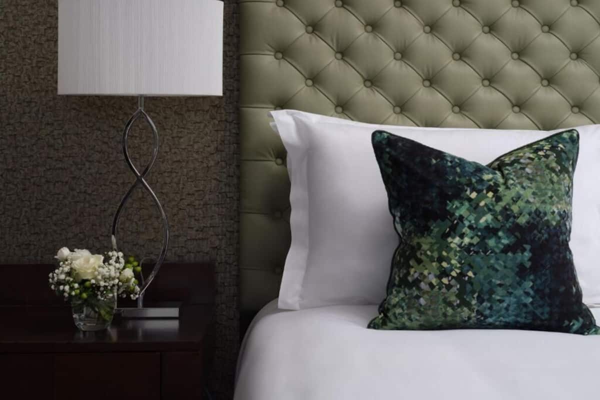 Ein Schlafzimmer mit einer gesteppten Kopfteil, einem weißen Kissen, einem Zierkissen, einem Nachttisch mit Blumen in einer Vase und einer Wandleuchte.