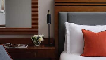 A hotel room with a wooden headboard, a bedside table with a lamp, a bed with white and orange pillows, and a small vase with flowers.