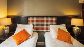Habitación de hotel con dos camas individuales, cada una con sábanas blancas y almohadas naranjas. Hay dos lámparas de noche con pantallas, y un cabecero a cuadros sobre las camas.