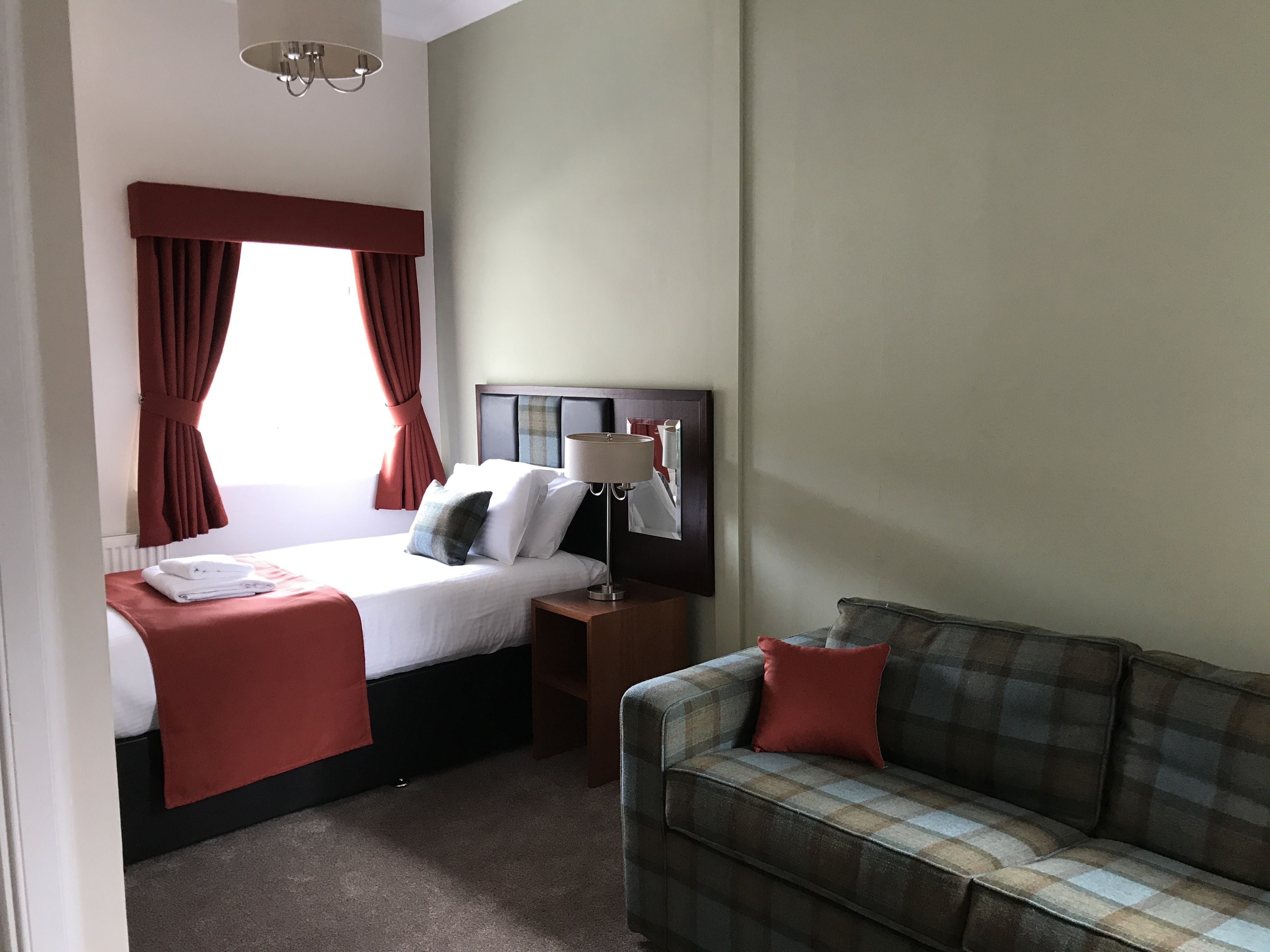 Una habitación de hotel con cama, mesita de noche, lámpara, sofá y ventana con cortinas.
