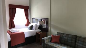 Una habitación de hotel con cama, mesita de noche, lámpara, sofá y ventana con cortinas.