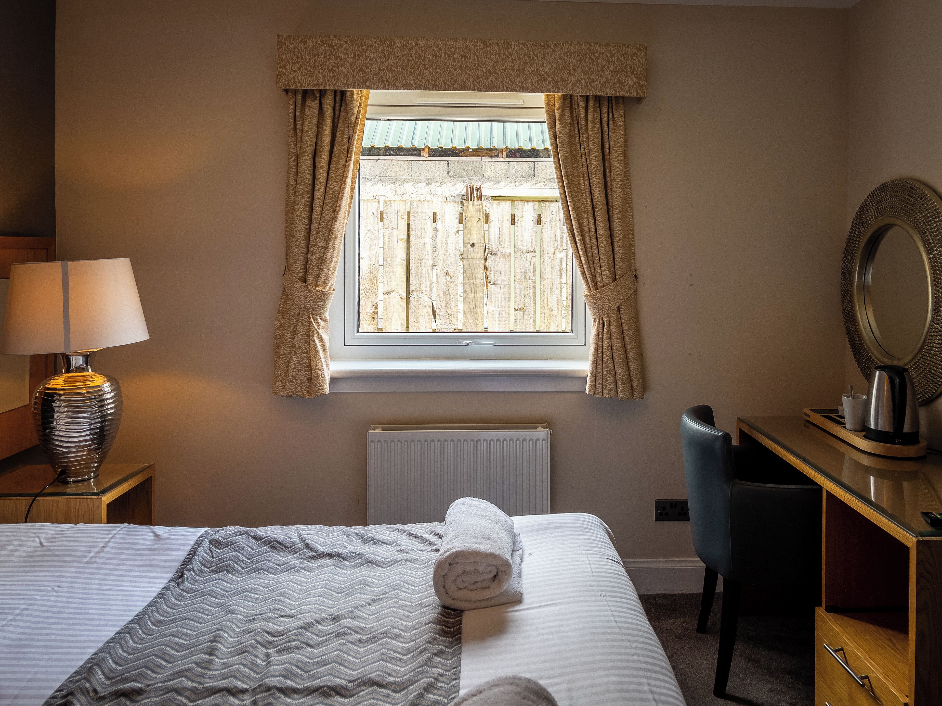Hotelkamer met een bed, bureau, stoel, lamp, radiator en raam met gordijnen.