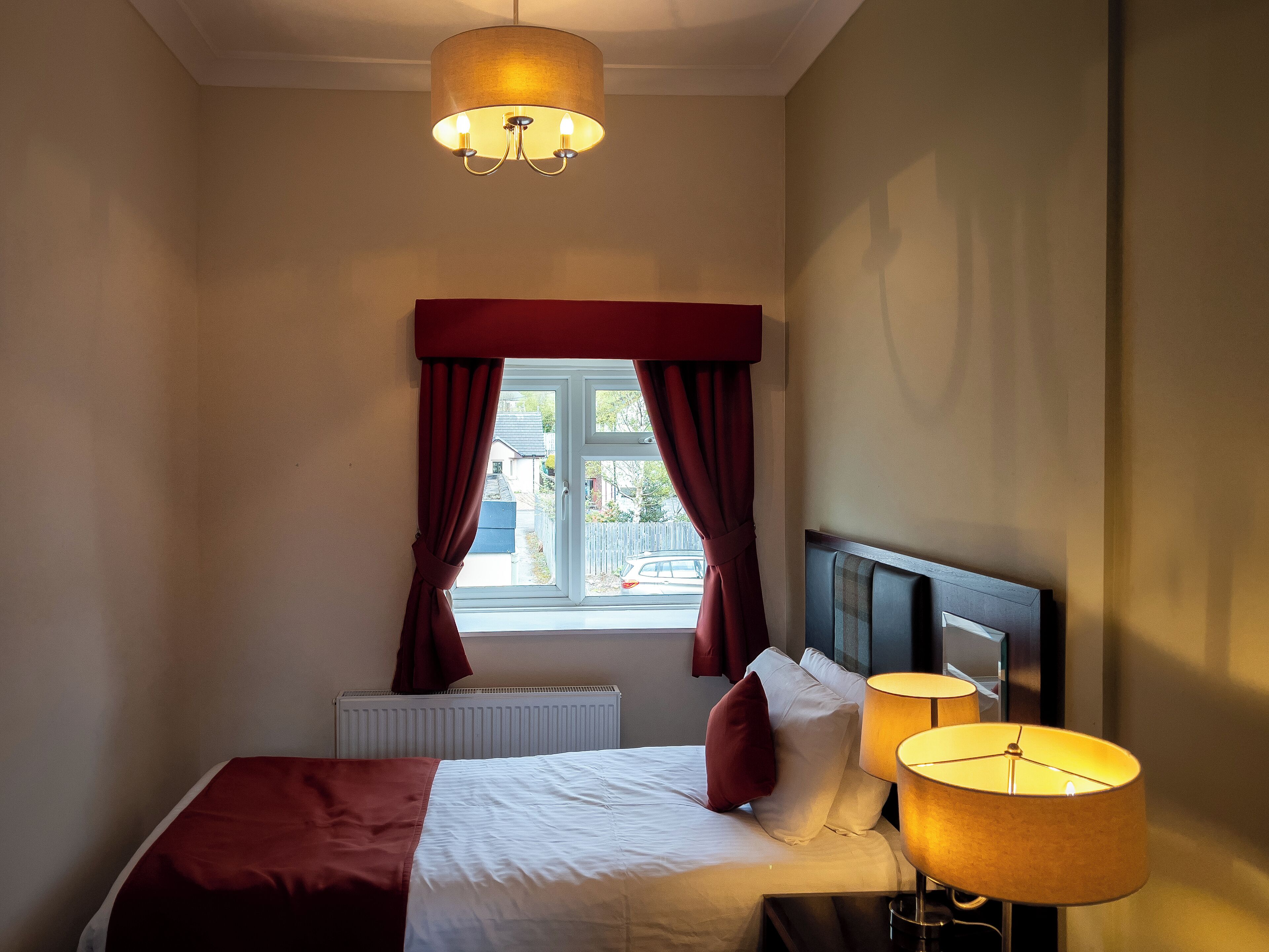 Een hotelkamer met een bed, een raam met gordijnen, een radiator en een bedlamp.