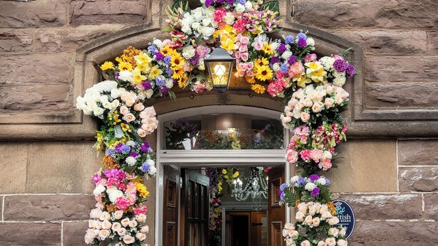 En entré till en byggnad av sten, med ett rådjursskelett ovanför, en färgstark blomsterbåge och skyltar med texten ”DESH Restaurant” och ”CRAGS BAR – öppnar snart”.