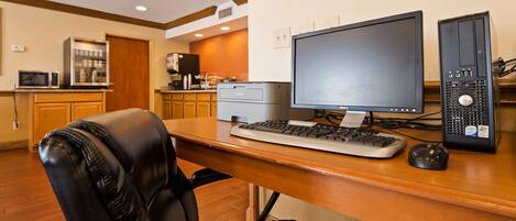 A hotel room with a wooden desk, a black leather chair, a Dell monitor, a desktop computer, and a Brother printer.