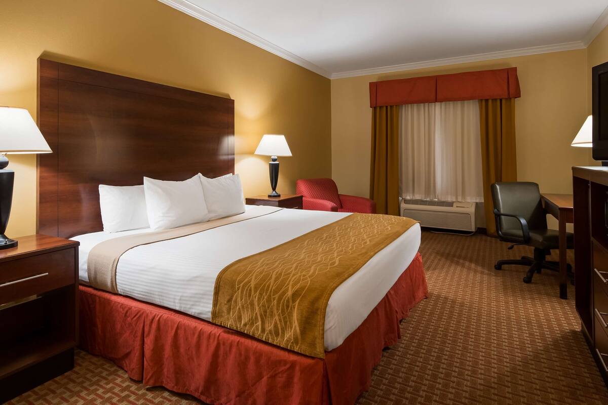 A hotel room with a large bed, two bedside tables with lamps, a desk with a chair, a television, and an air conditioning unit.