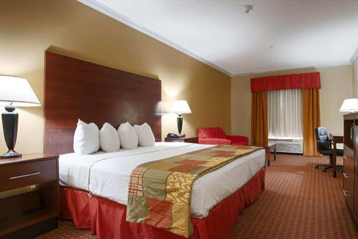 A hotel room with a large bed, two bedside tables with lamps, a red chair, a desk with a computer, and a TV on a stand.