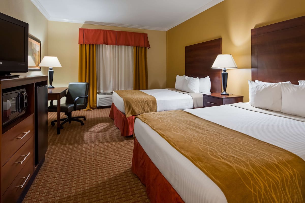 A hotel room with two beds, a desk, a chair, a TV, and a lamp.