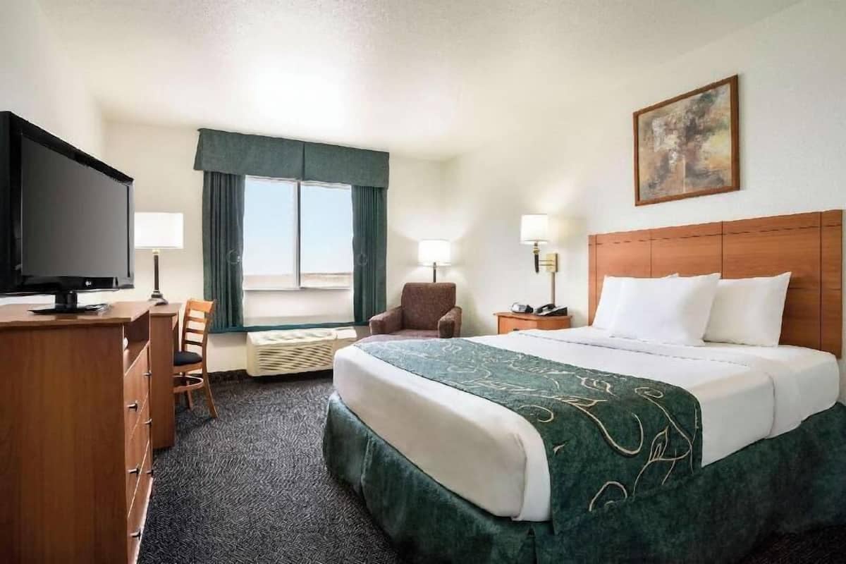 Ein Hotelzimmer mit einem Bett, einem Schreibtisch mit Fernseher, einem Sessel, einem Nachttisch, einer Lampe und einem Fenster mit Vorhängen.