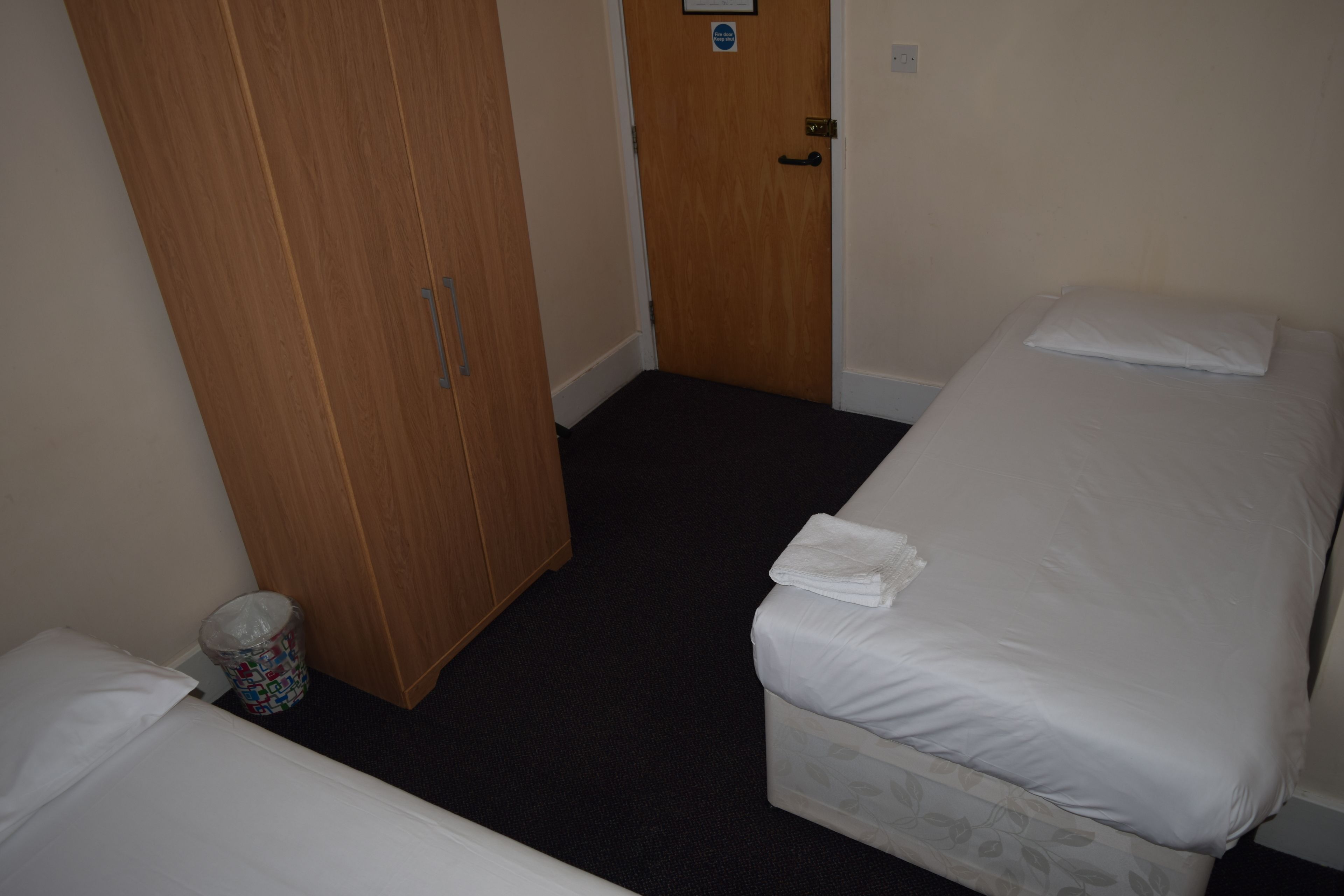 A small, single-room hotel room with a wooden wardrobe, a small trash bin, and a bed with a white pillow and a folded towel.