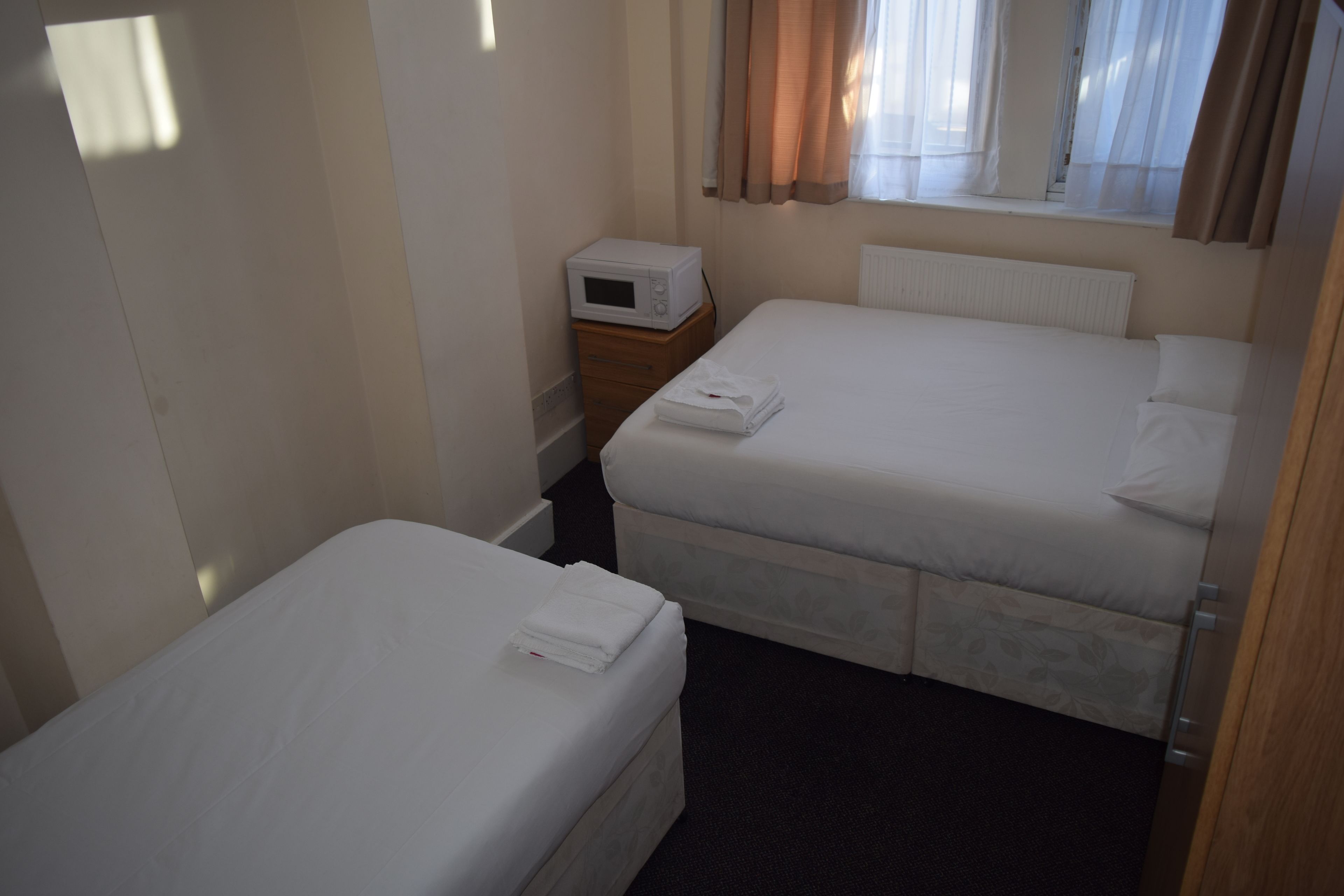 Ein kleines, sauberes Zimmer mit zwei Einzelbetten, einem Nachttisch, einer Mikrowelle und einem Fenster mit Vorhängen.