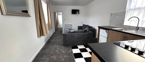 A modern kitchen with a black countertop and a sink, a grey sofa in the living area, and a checkered floor.