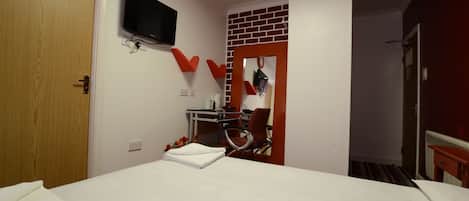 Habitación de hotel con cama, escritorio con silla, televisor y pared de ladrillo rojo como detalle decorativo.