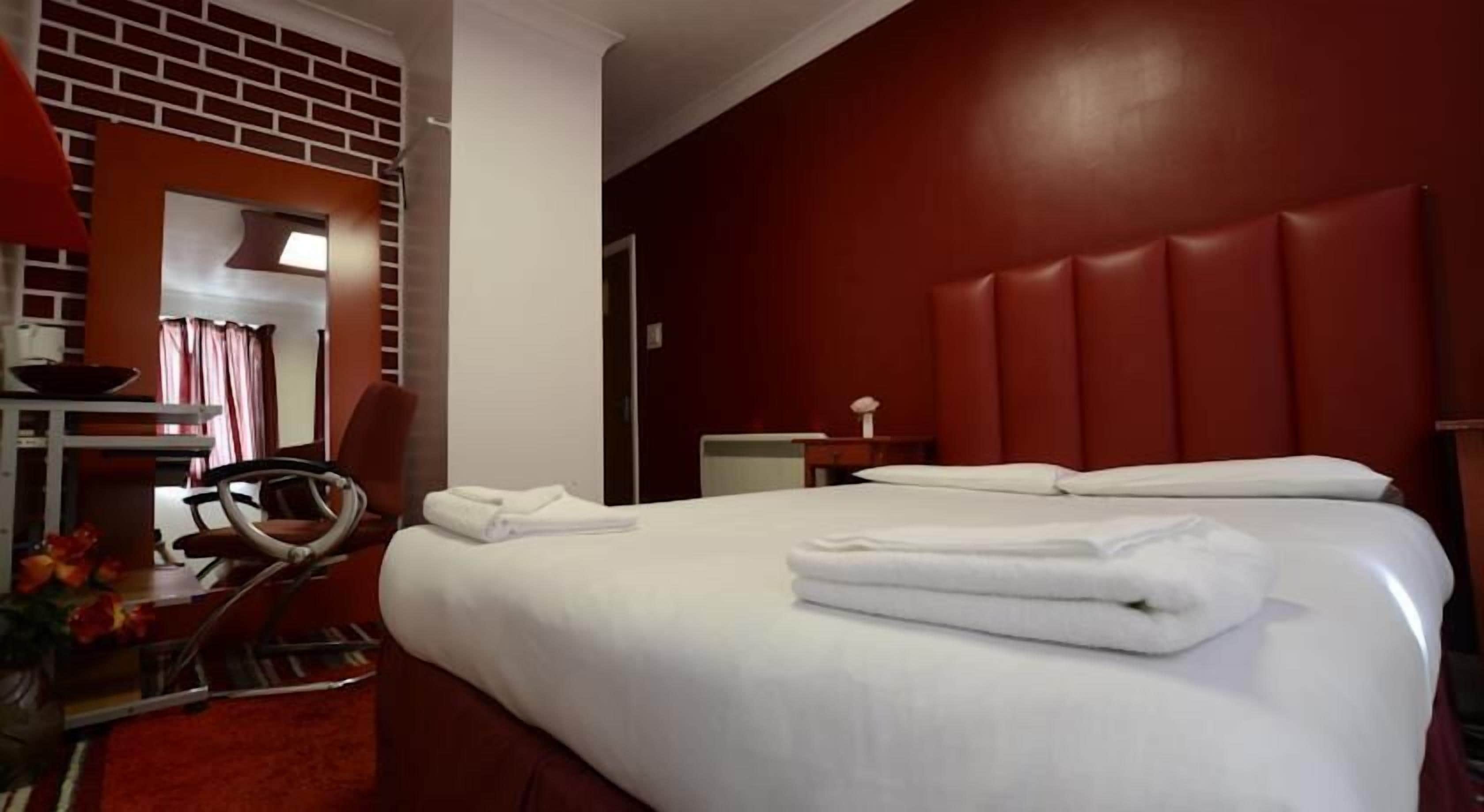 A hotel room with a large bed, a desk with a chair, a mirror, and a red brick wall.