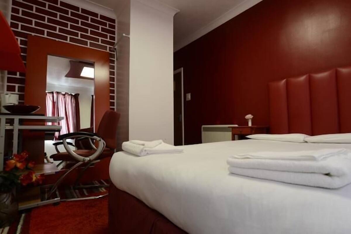 A hotel room with a large bed, a desk with a chair, a mirror, and a red brick wall.