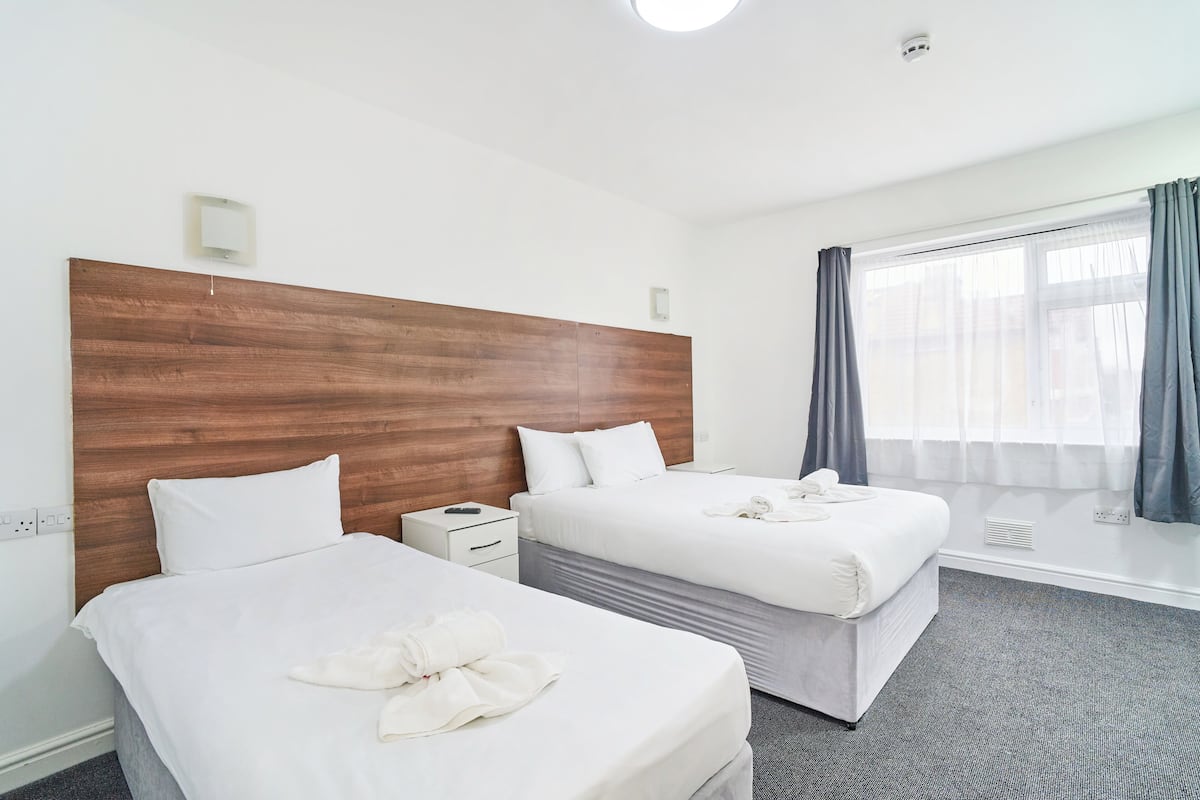 Habitación de hotel con dos camas, cabecero de madera, mesita de noche y un amplio ventanal con cortinas.