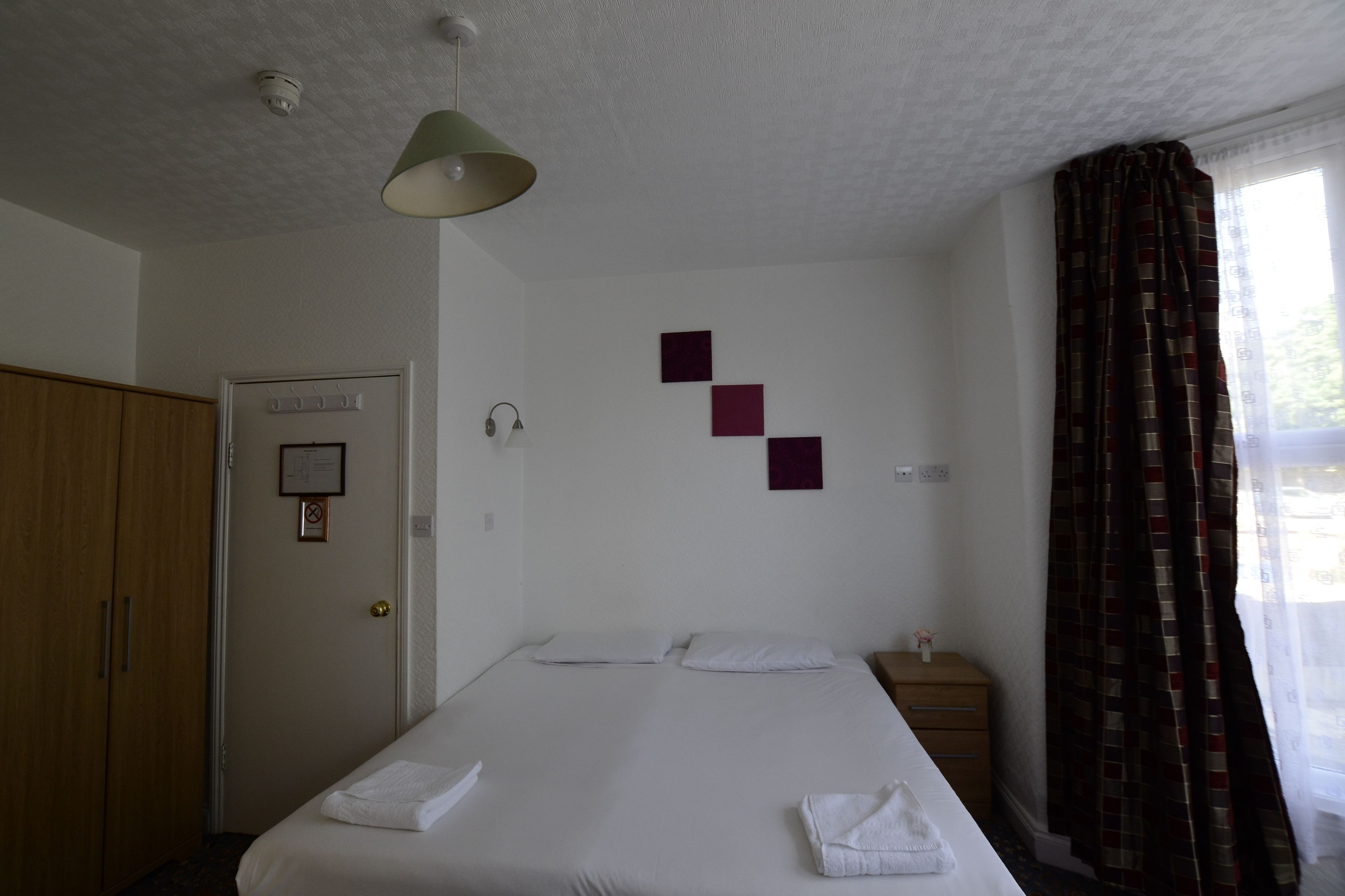 A hotel room with a bed, a wardrobe, a bedside table, and a window with curtains.