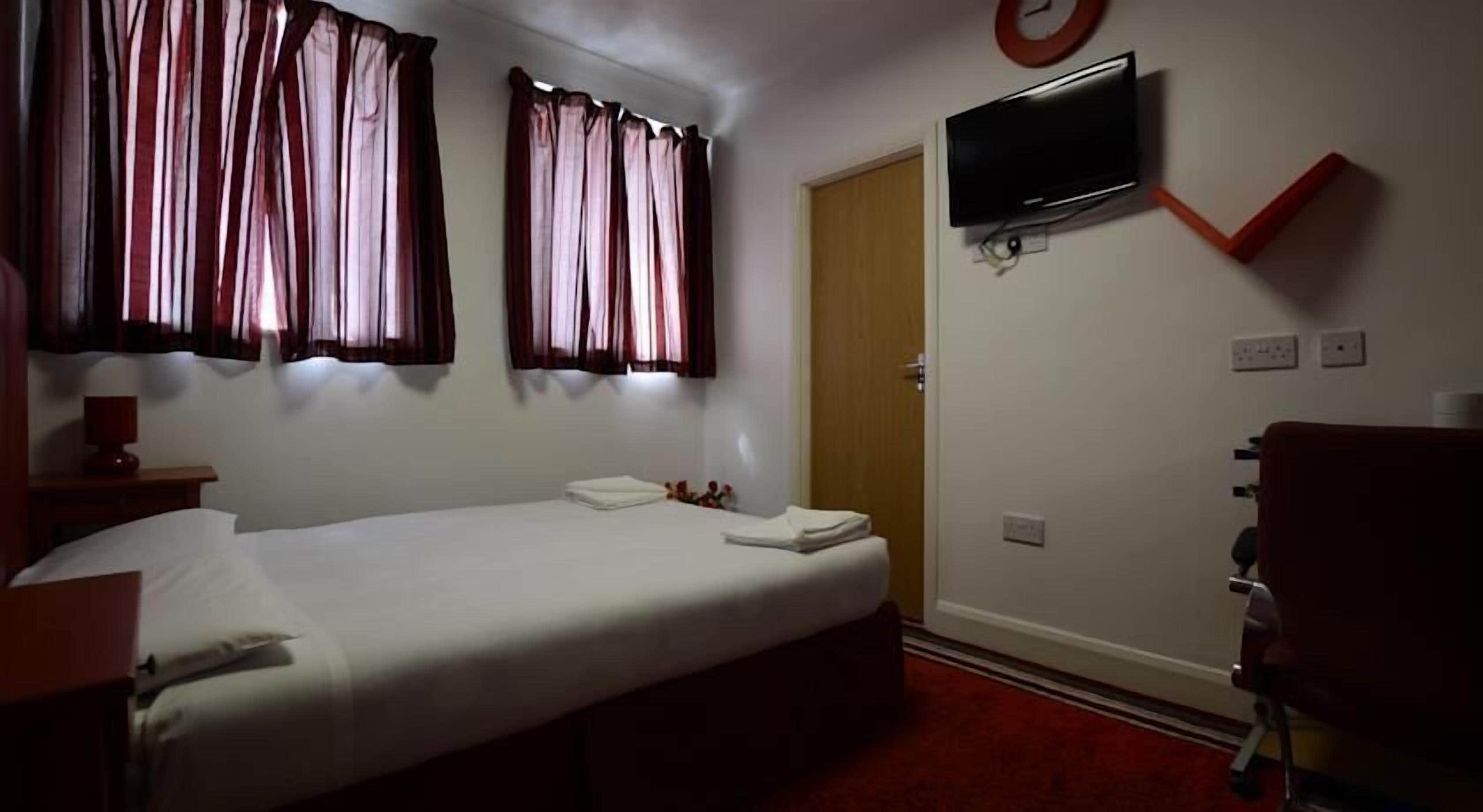A single bed with white linens, a bedside table with a lamp, a television mounted on the wall, and a red carpeted floor.
