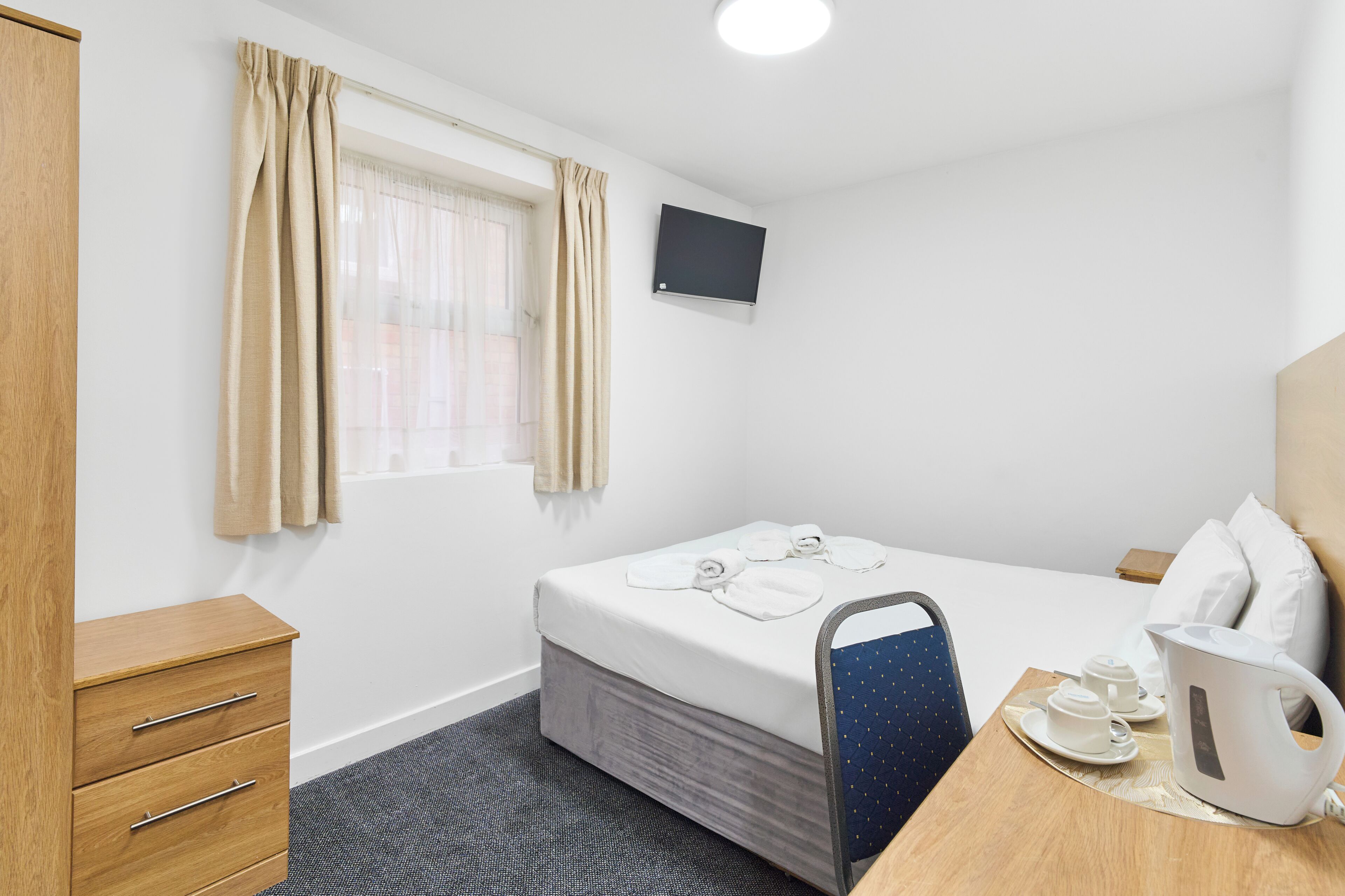 A hotel room with a bed, a desk with a kettle, a chair, a TV, and a wardrobe.