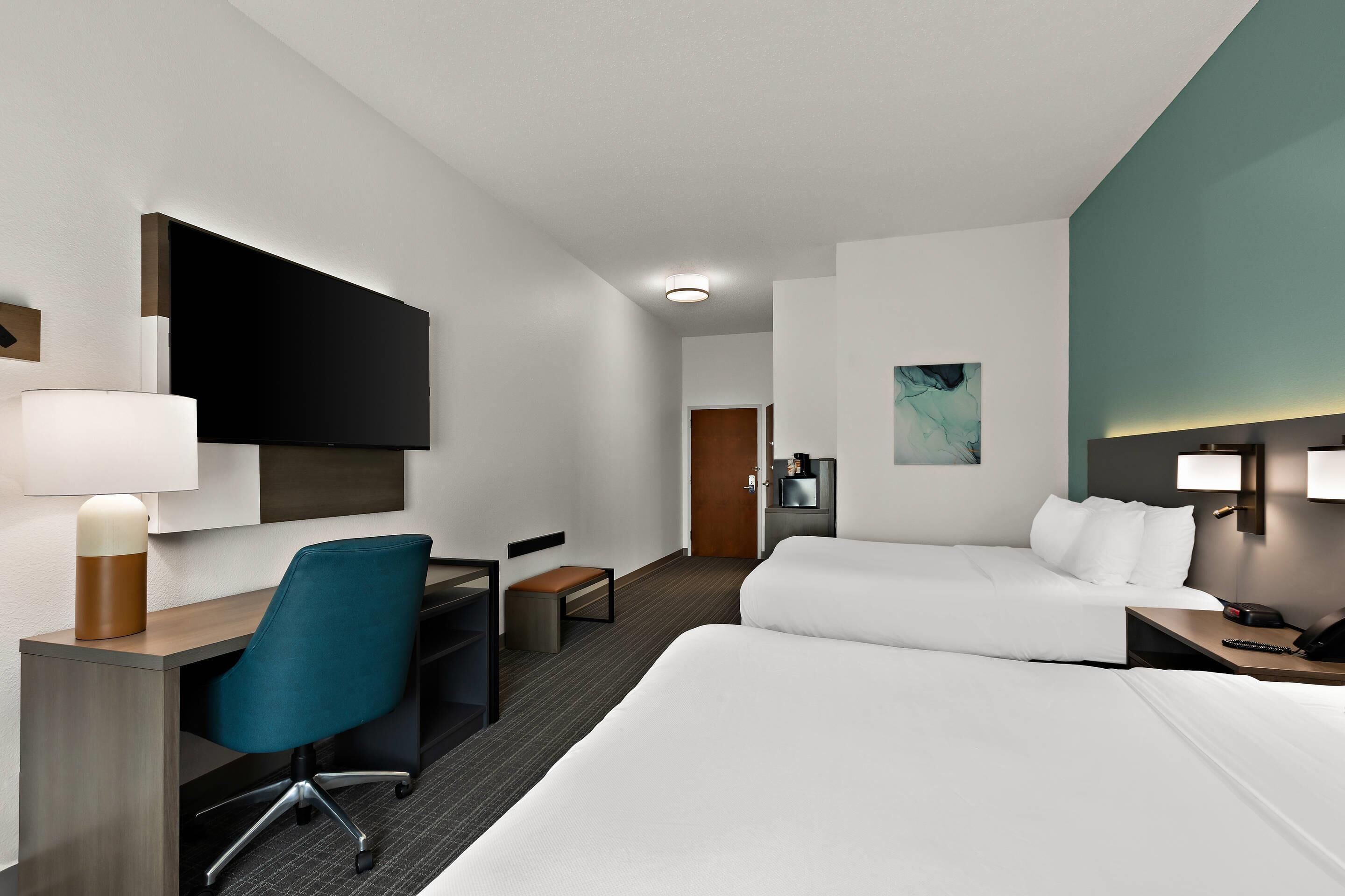 A hotel room with a bed, a desk with a chair, a TV, a small table, and a nightstand with a lamp.