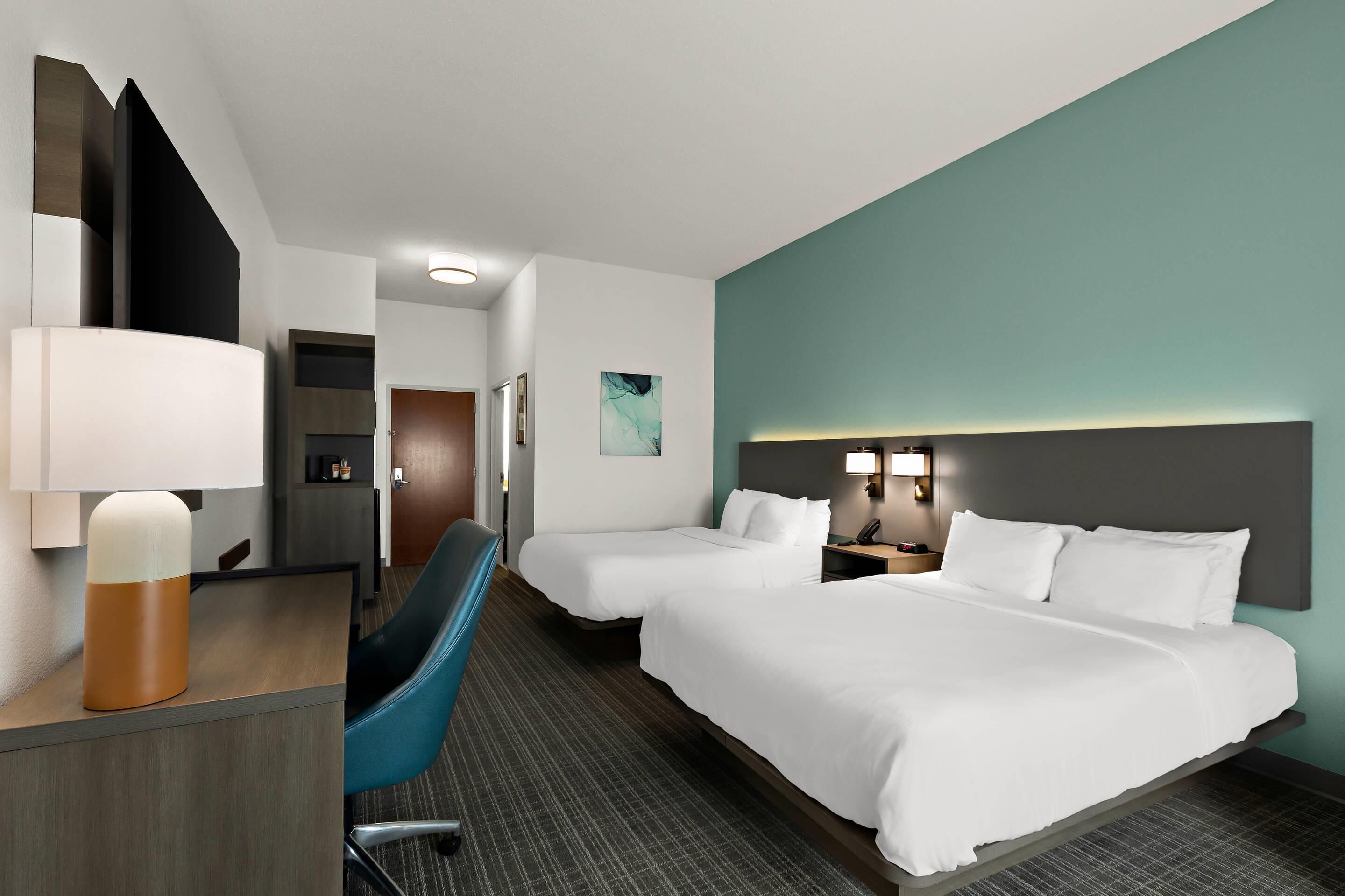 Habitación de hotel con dos camas, un escritorio con lámpara, una silla y un baño a la vista en el fondo.
