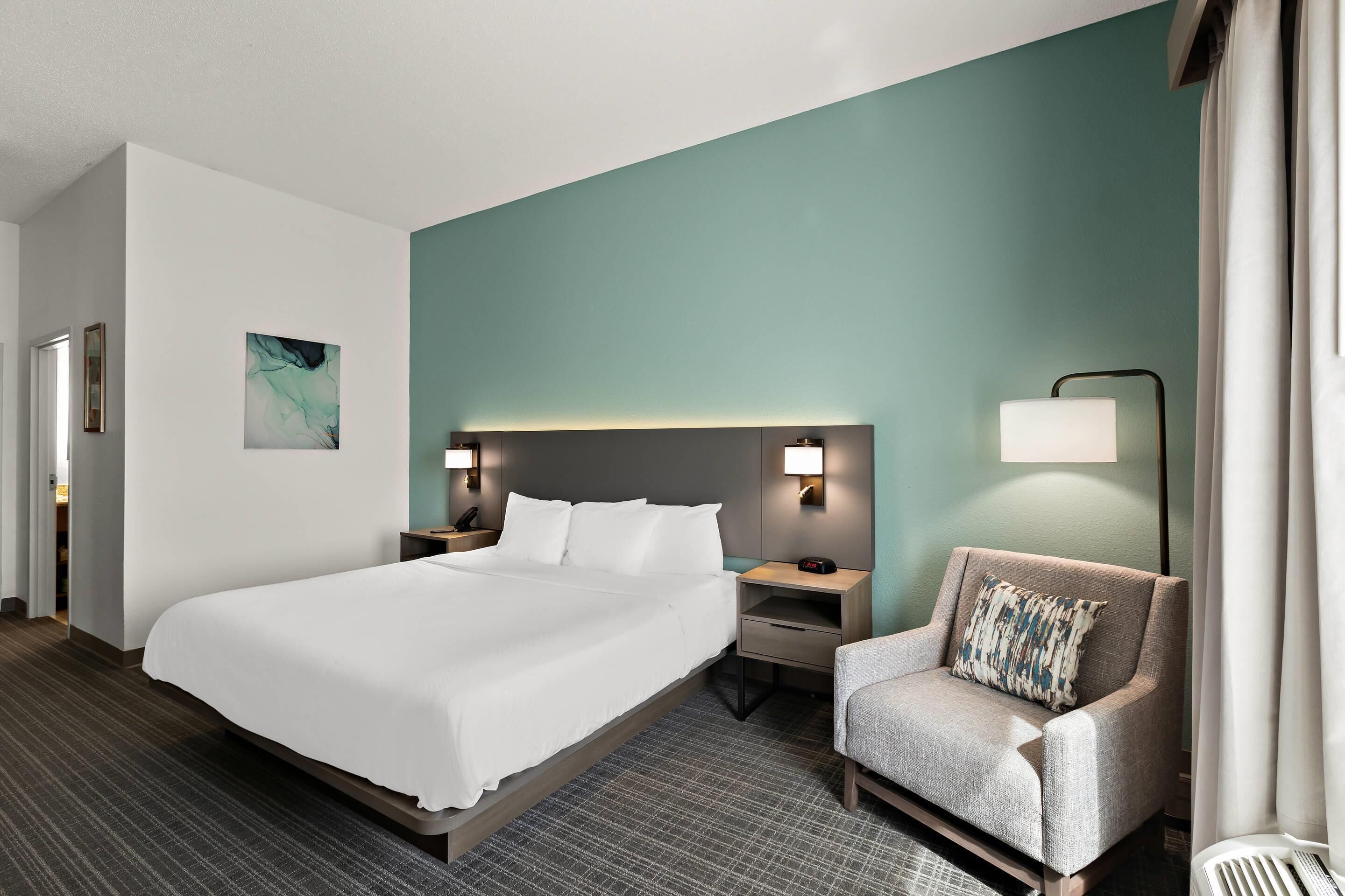 Una habitación de hotel con una cama grande, una mesita de noche, una silla y una lámpara fijada a la pared.