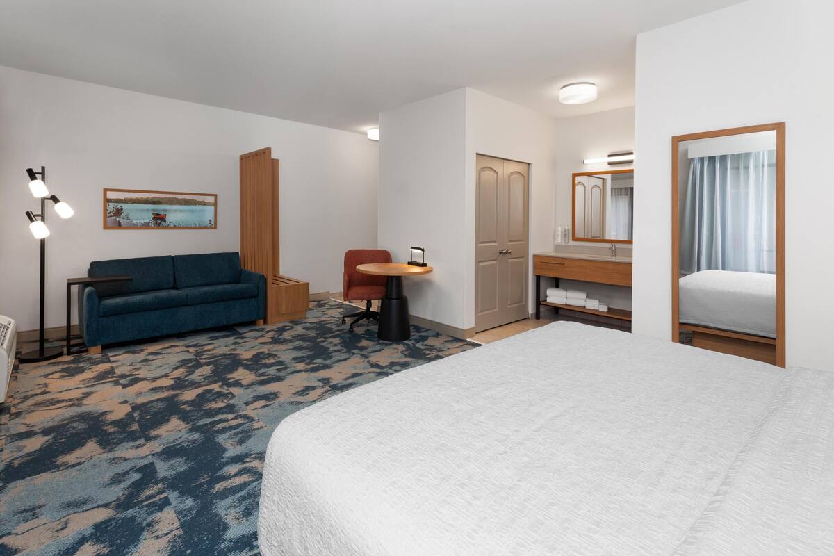 Quarto de hotel com cama, sofá, escrivaninha, cadeira, guarda-roupa e espelho.