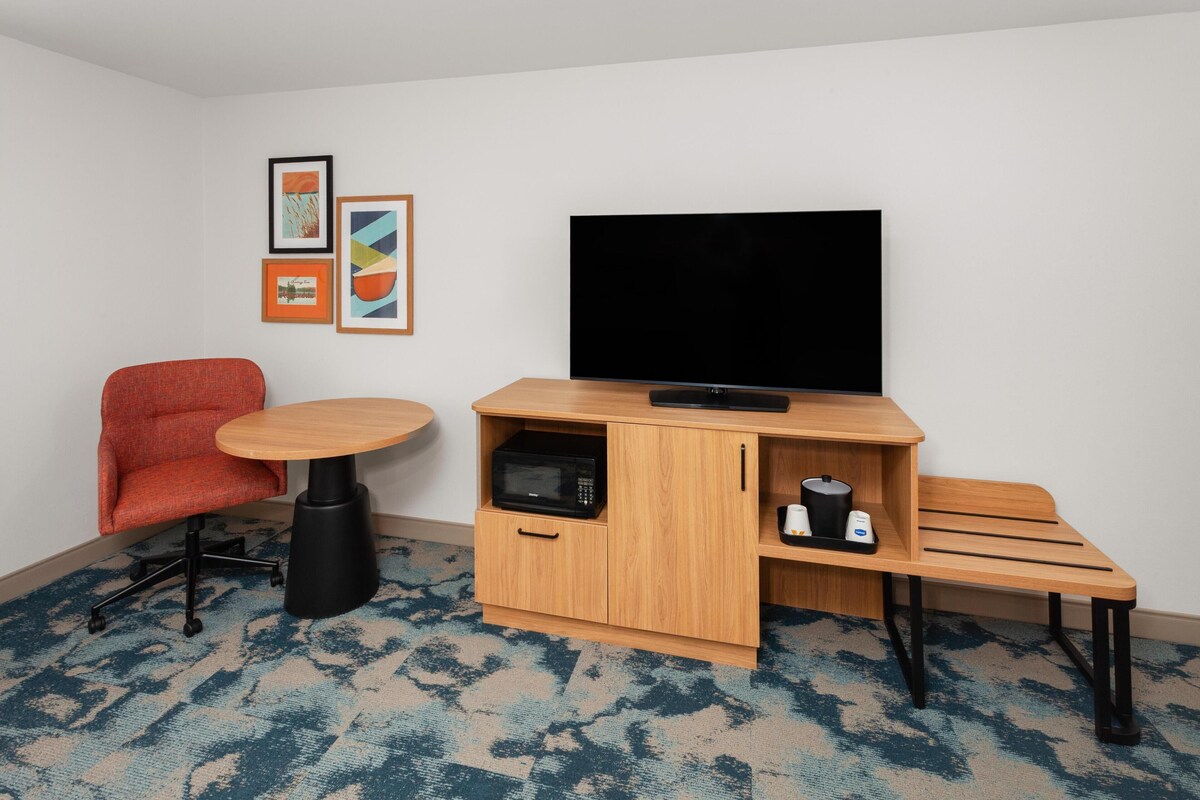 A compact room with a single red chair, a small round table, a television on a wooden stand, a microwave, and a bench.