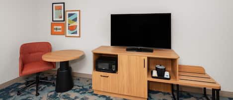 A compact room with a single red chair, a small round table, a television on a wooden stand, a microwave, and a bench.