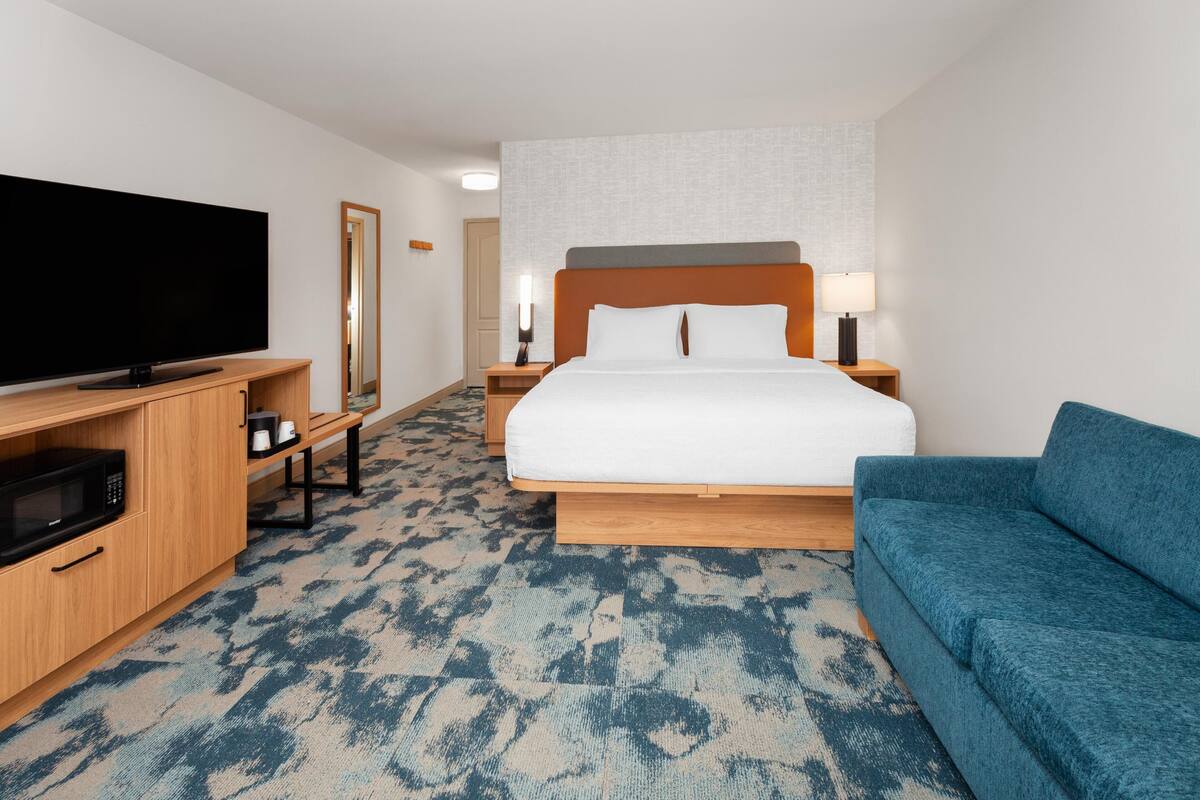 Quarto de hotel com cama, televisão, micro-ondas, sofá e piso coberto por carpete.
