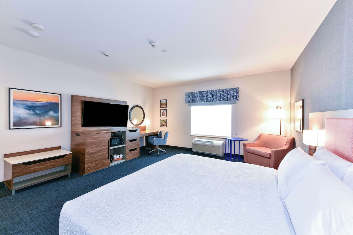 A hotel room with a large bed, a desk with a computer, a TV, a chair, and a window with blinds.