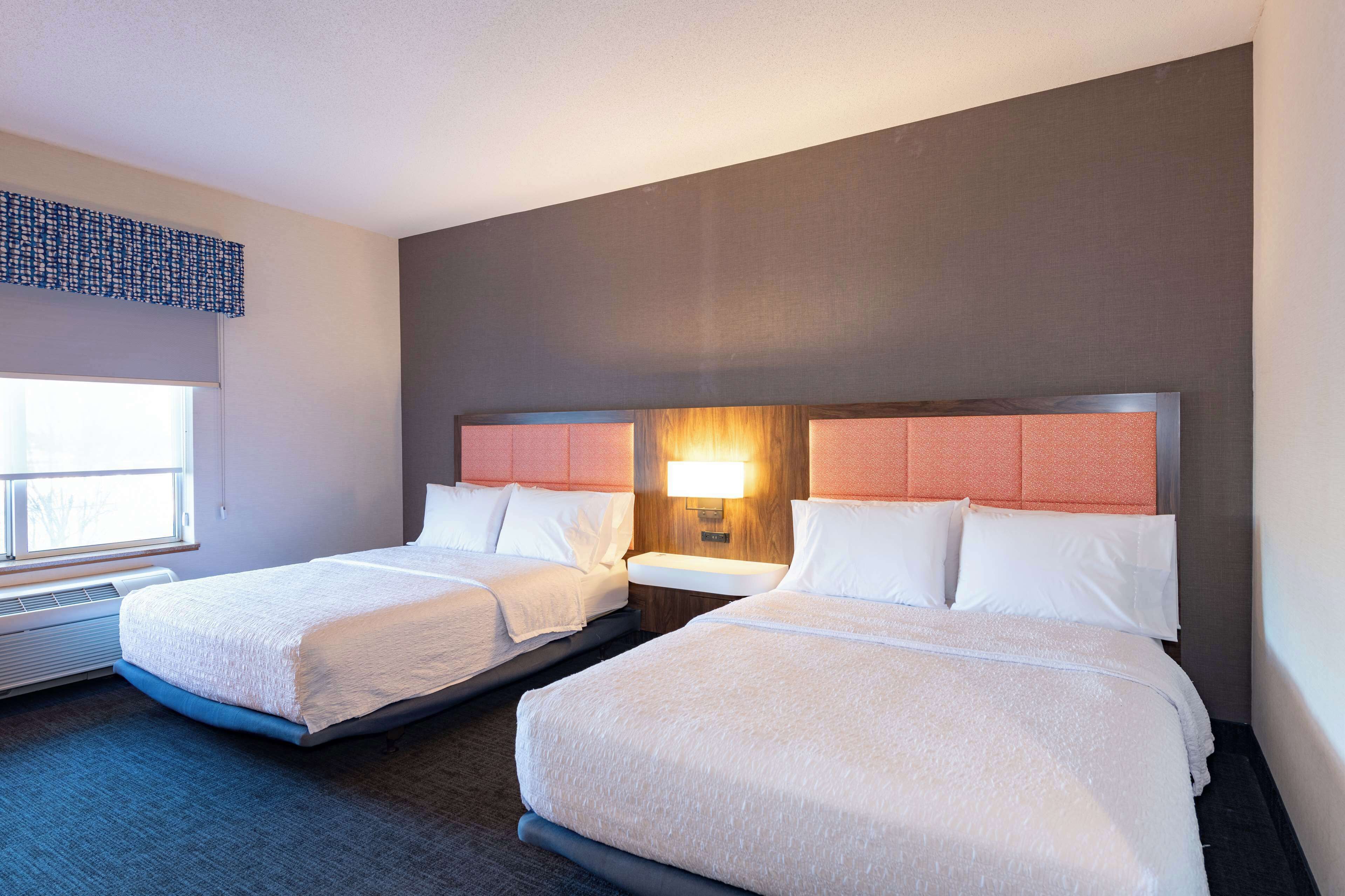 A hotel room with two beds, a window with blinds, a wall-mounted lamp, and a heater.