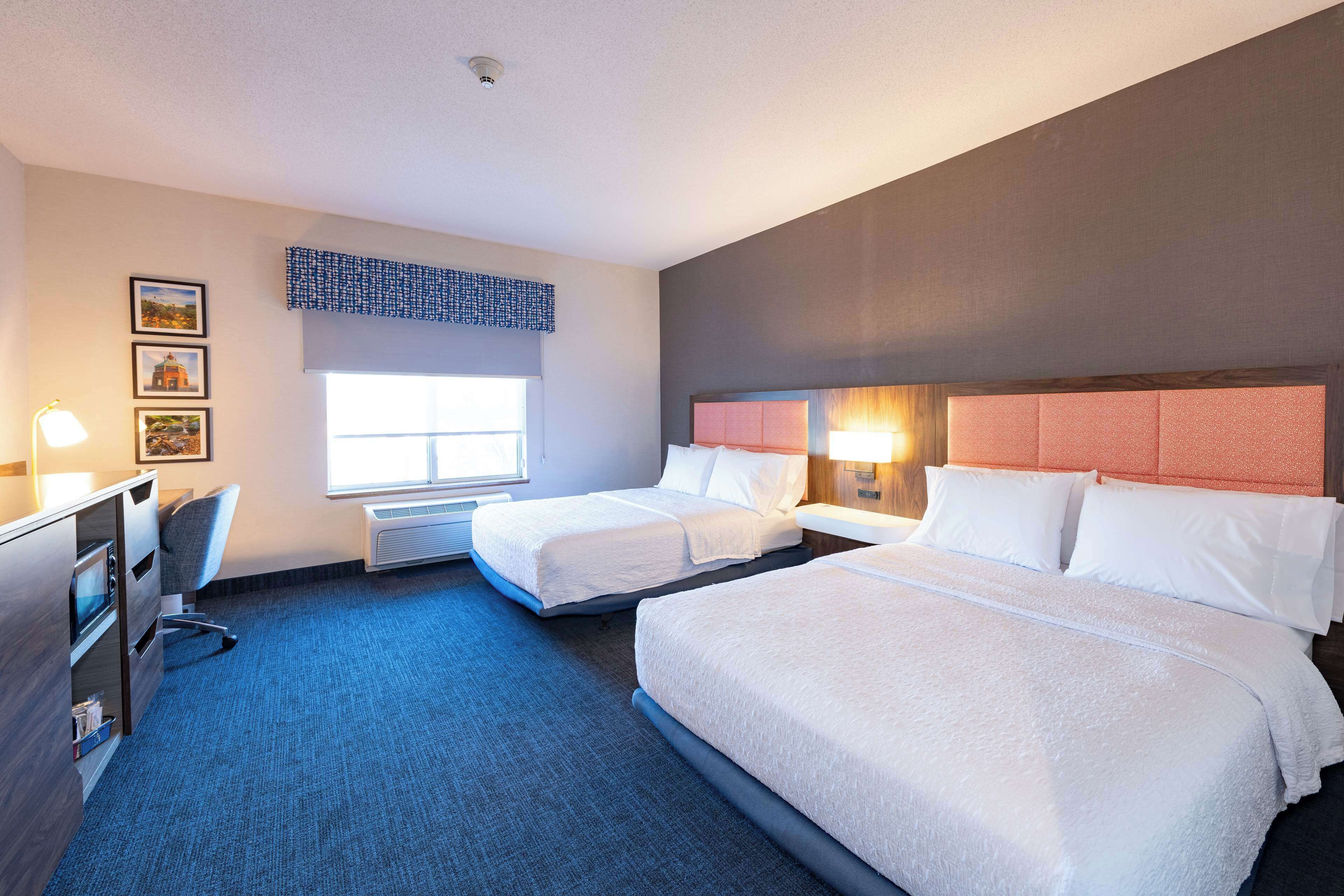 A hotel room with two beds, a desk, a chair, a window, and a wall-mounted air conditioner.
