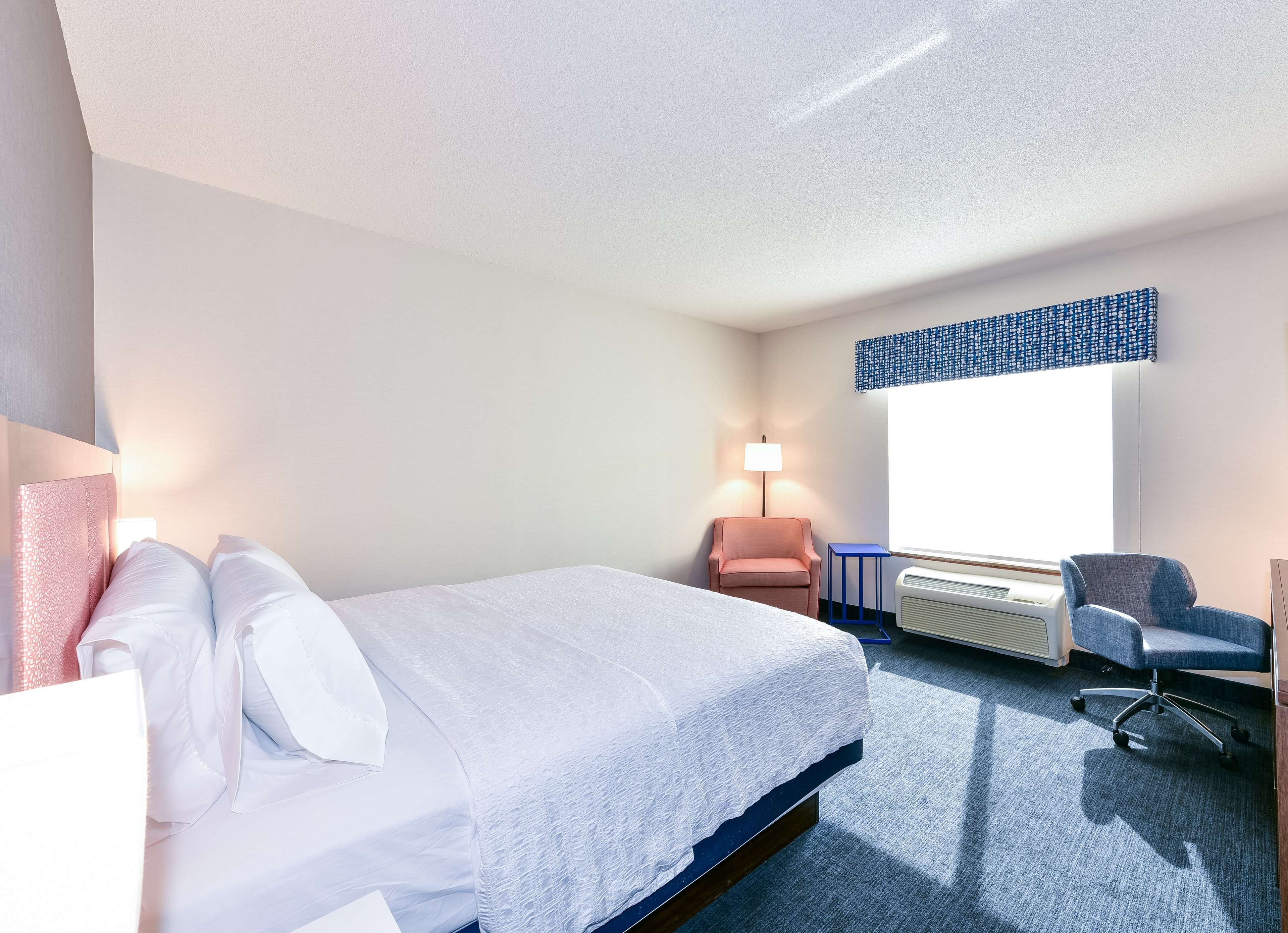 A hotel room with a bed, a desk, a chair, a lamp, and a window.