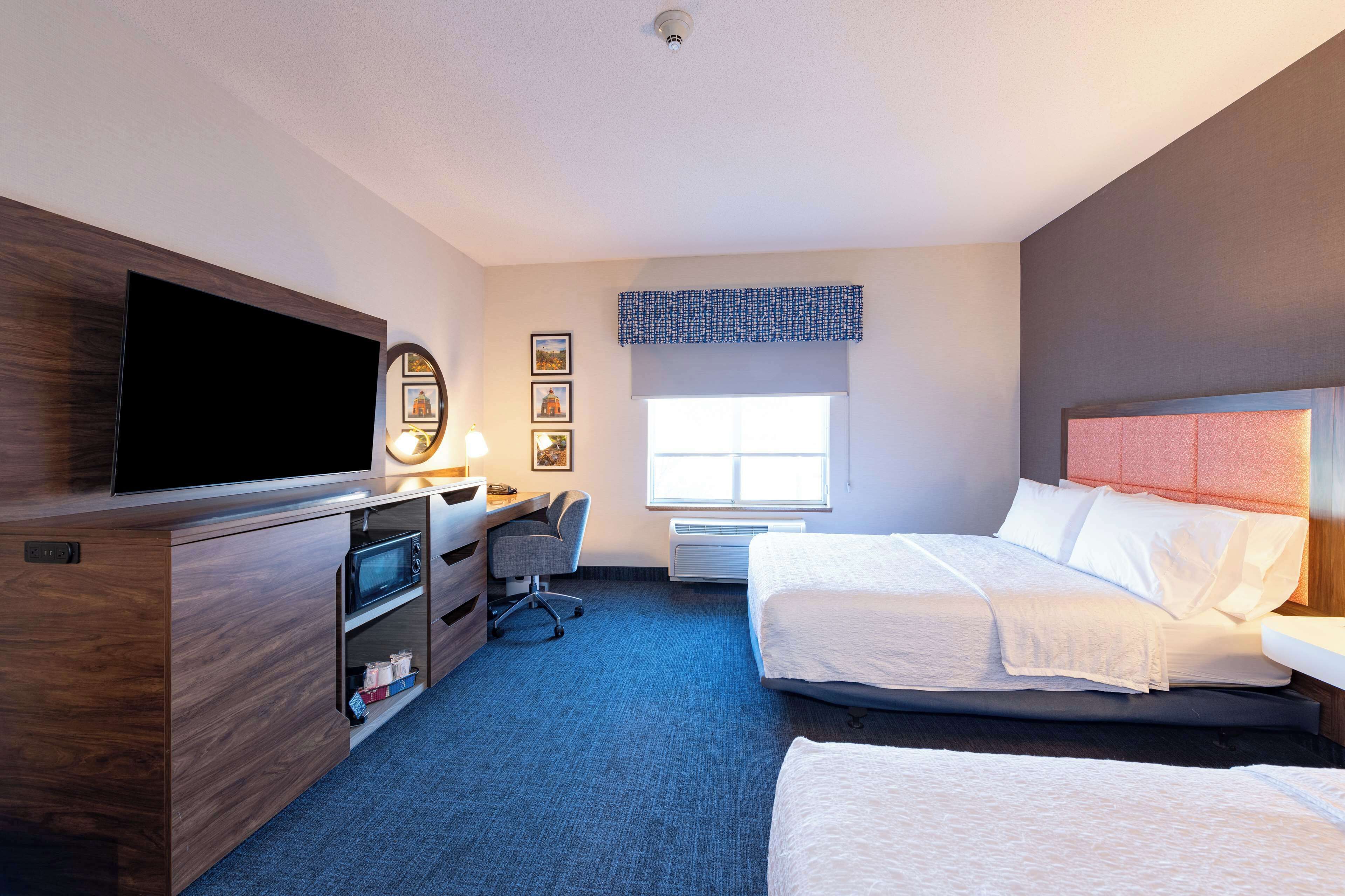 A hotel room with a flat-screen TV, a desk with a chair, two beds, a window with a view, and a ceiling-mounted light.