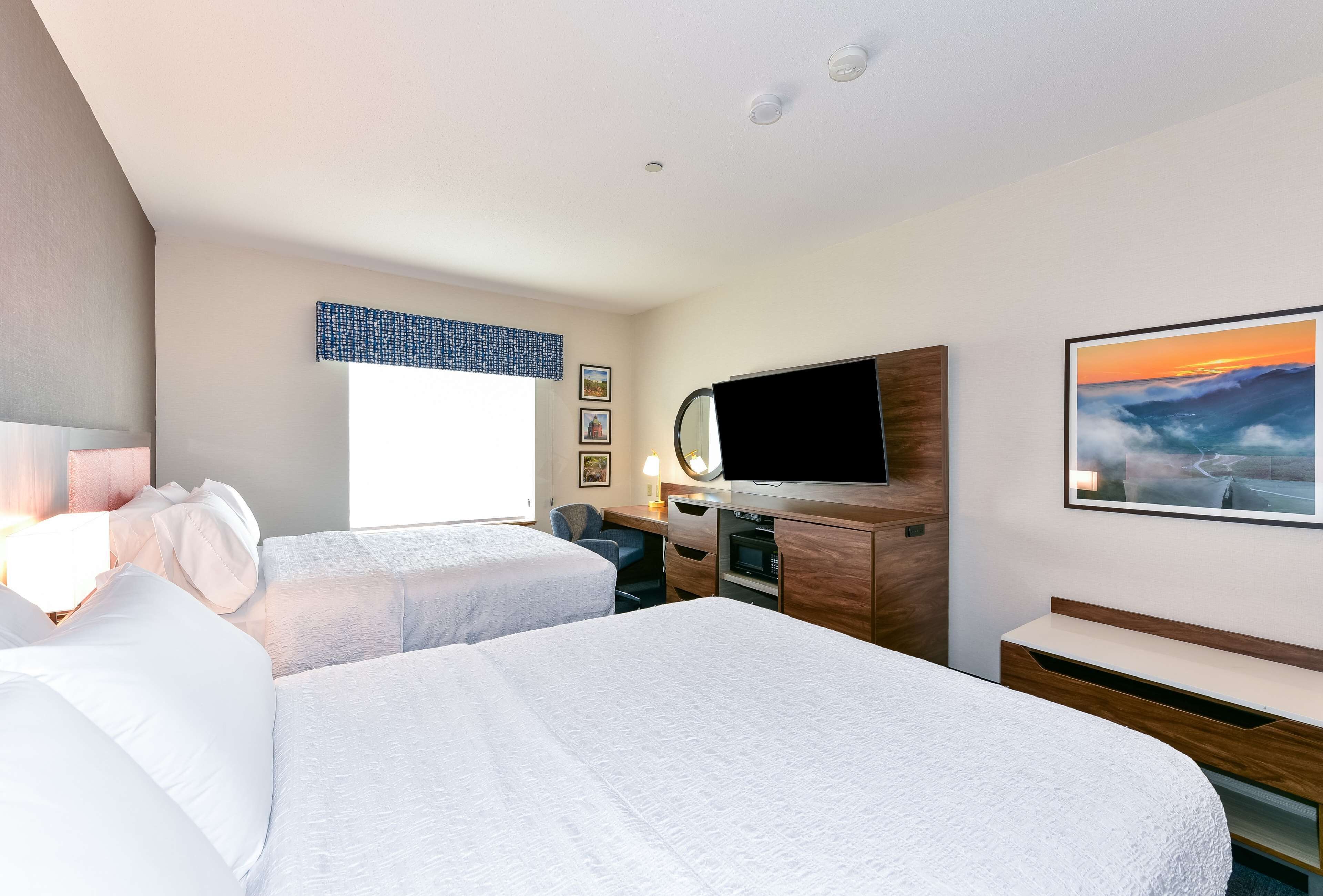 A hotel room with two beds, a desk, a TV, and a painting of a sunset.