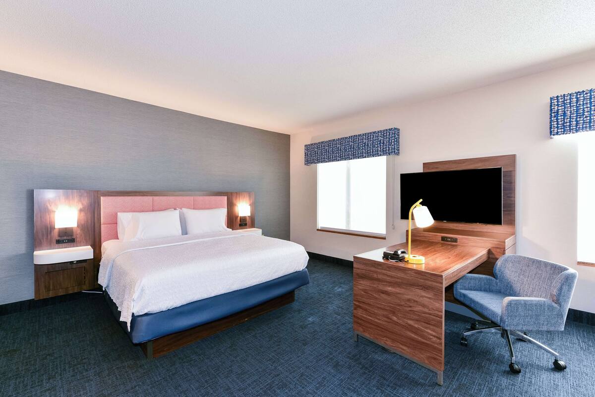 A hotel room with a bed, a desk, a chair, a TV, and a window with blinds.