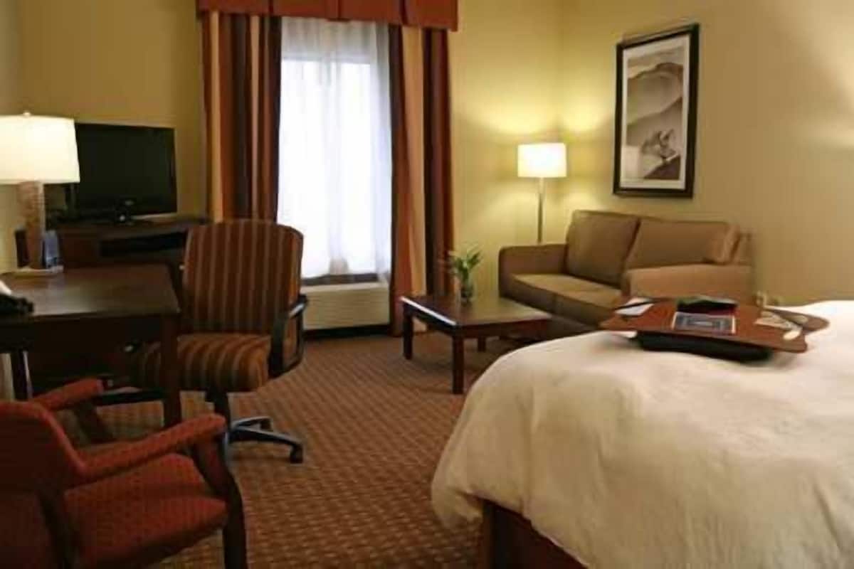 A hotel room with a bed, a desk with a lamp, a chair, a sofa, and a television.