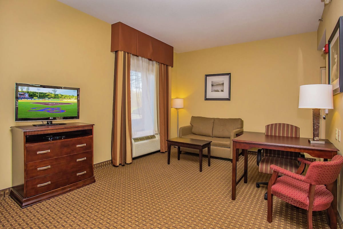 A hotel room with a flat-screen TV, a sofa, a dining table, a red chair, a lamp, a dresser, and a window with curtains.